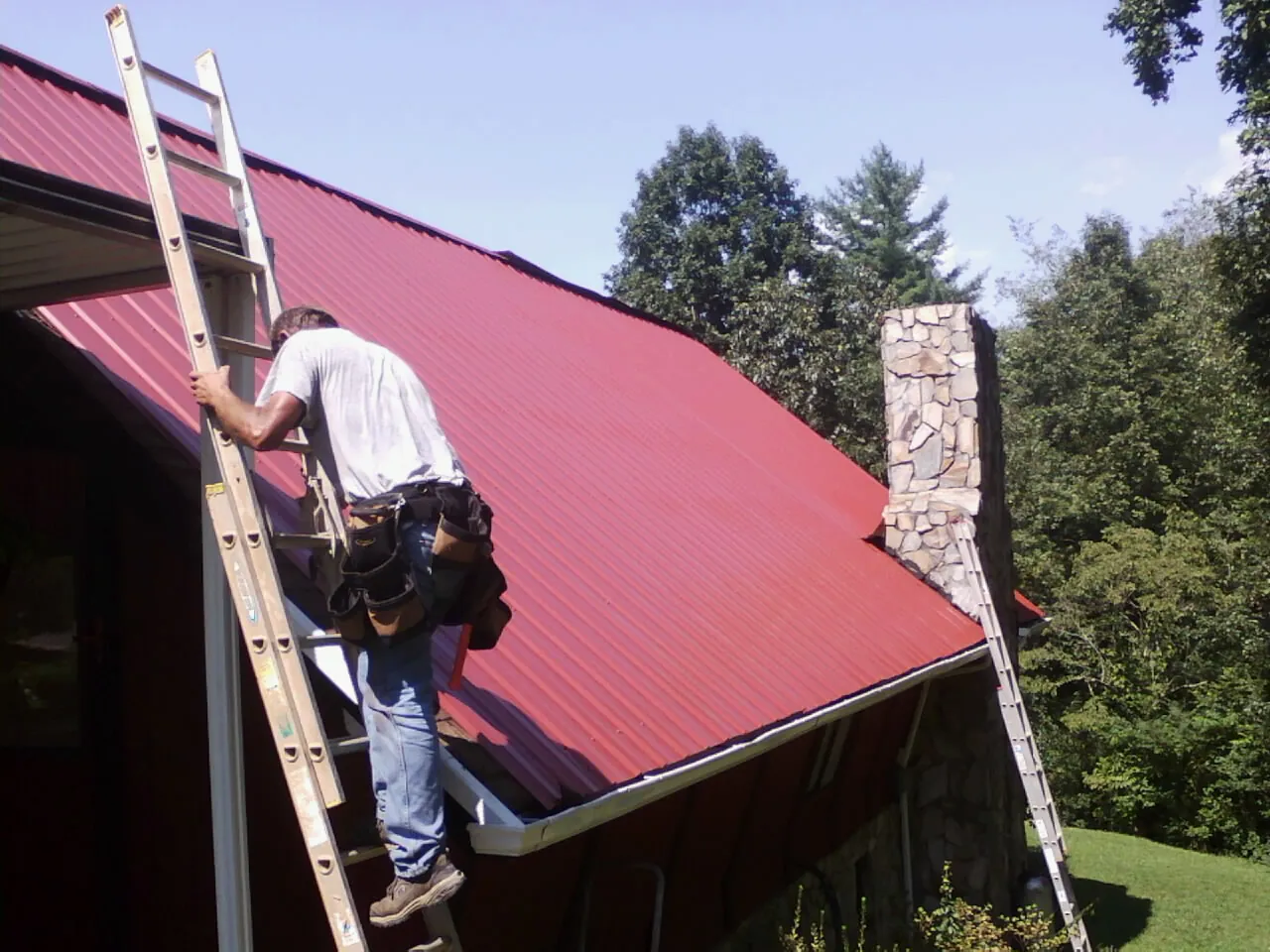 Roofer on a ladder beside a red metal roof and stone chimney — Soffit & Fascia Repair by Roofing Brothers in Uncasville